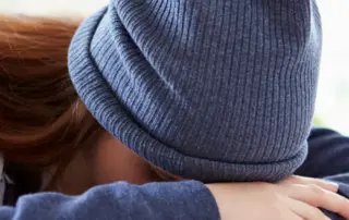 Woman Wearing Blue Hat With Hands On Forehead On Desk